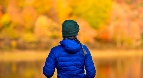 hiking and camping photo of woman in blud coat standing before a lake with fall foliage